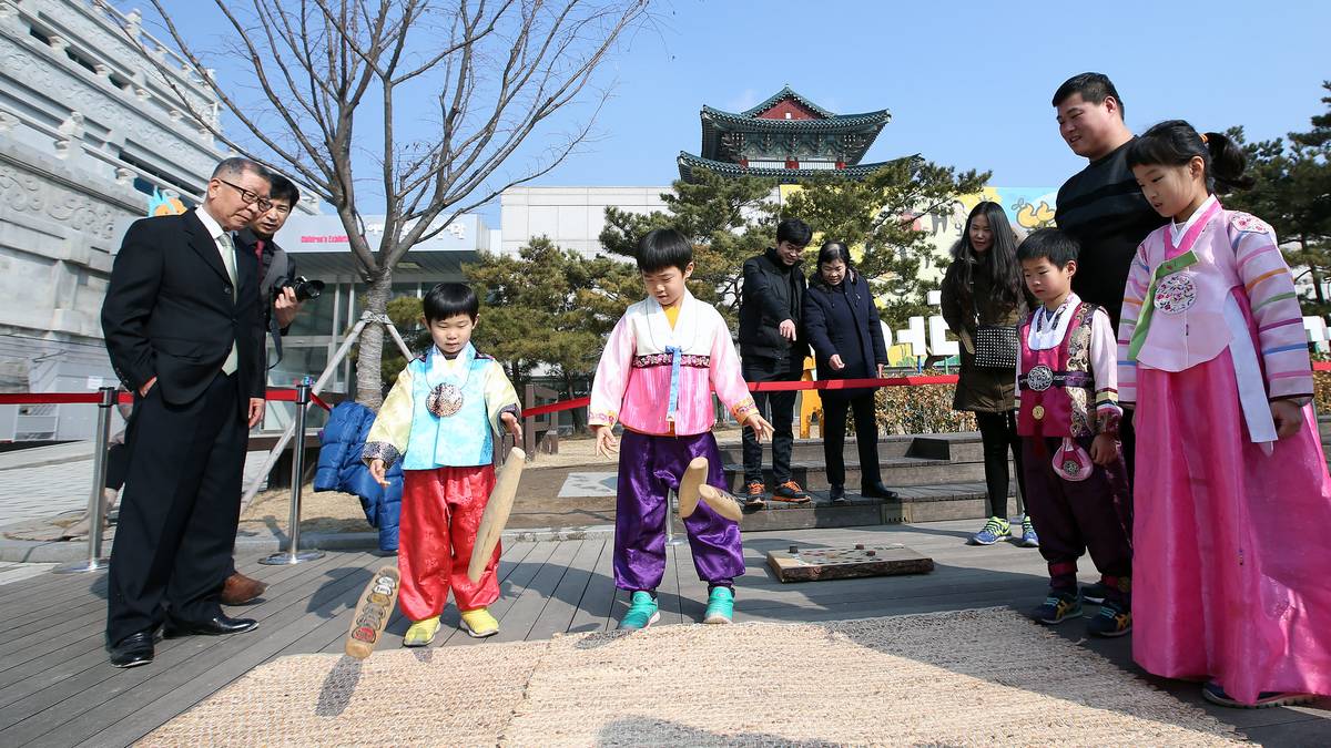Dois meninos de hanbok lançam varetas grandes de madeira sobre uma esteira ao ar livre, observados por adultos e outras crianças de hanbok