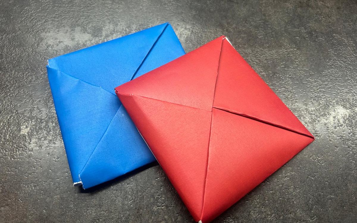 Dois quadrados de papel dobrado, um azul e um vermelho, sobre uma bancada escura