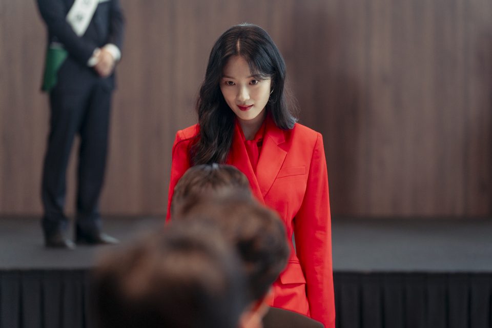 Kim Hye-yoon de terno vermelho diante de um palco de madeira, olhando para pessoas de costas em primeiro plano, com um homem de terno e faixa ao fundo
