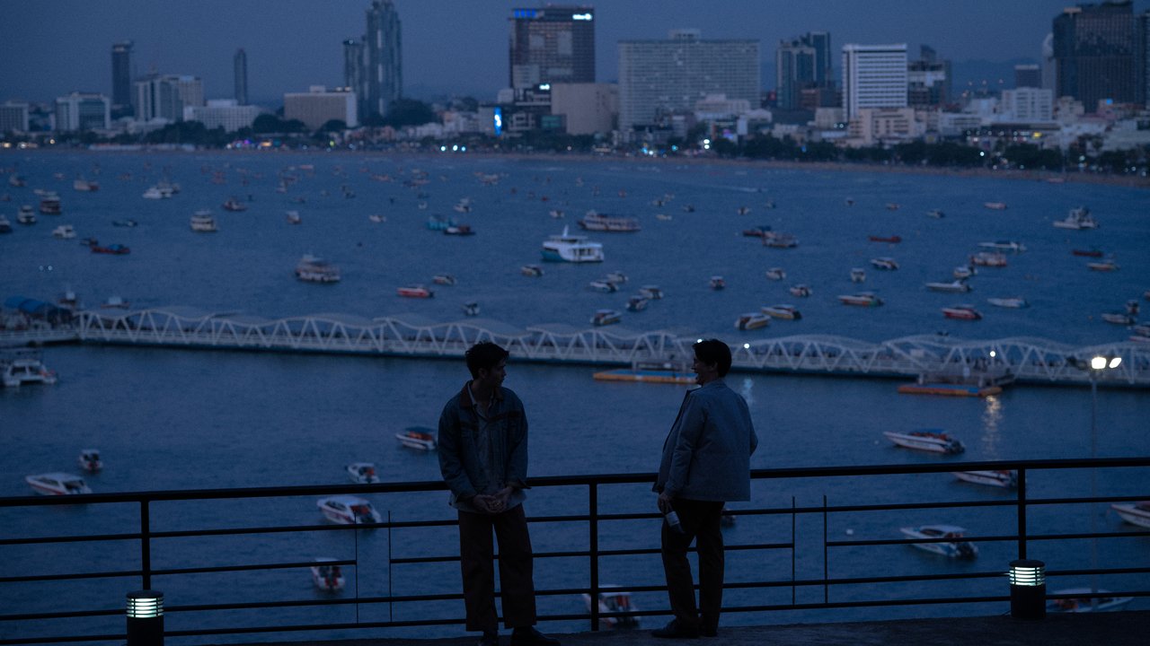 Dois homens de costas para a câmera junto a uma grade, diante de uma baía cheia de barcos e prédios iluminados ao entardecer