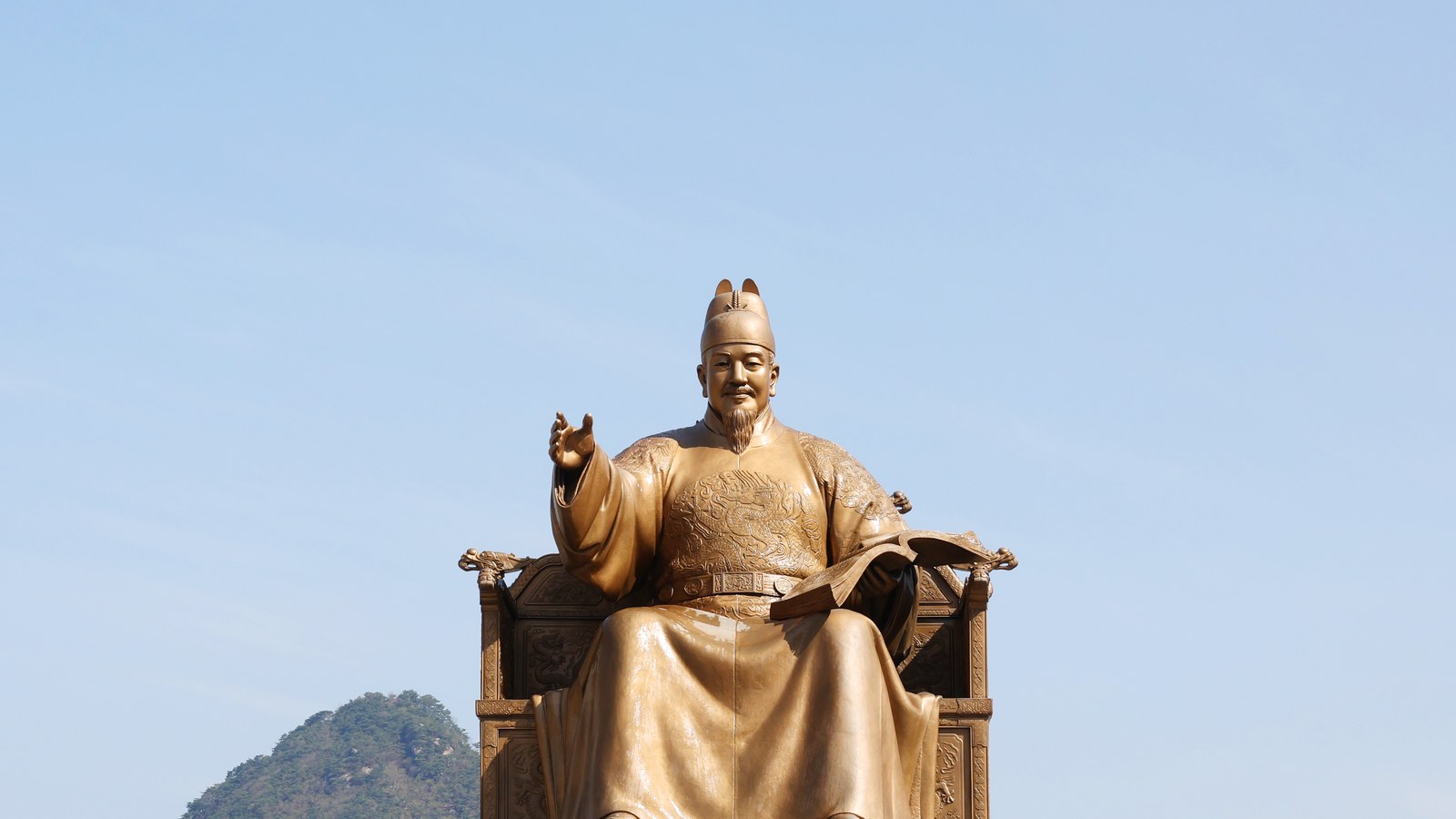 Estátua dourada do rei Sejong sentado no trono, com o braço direito estendido e um livro aberto sobre o colo, contra o céu azul