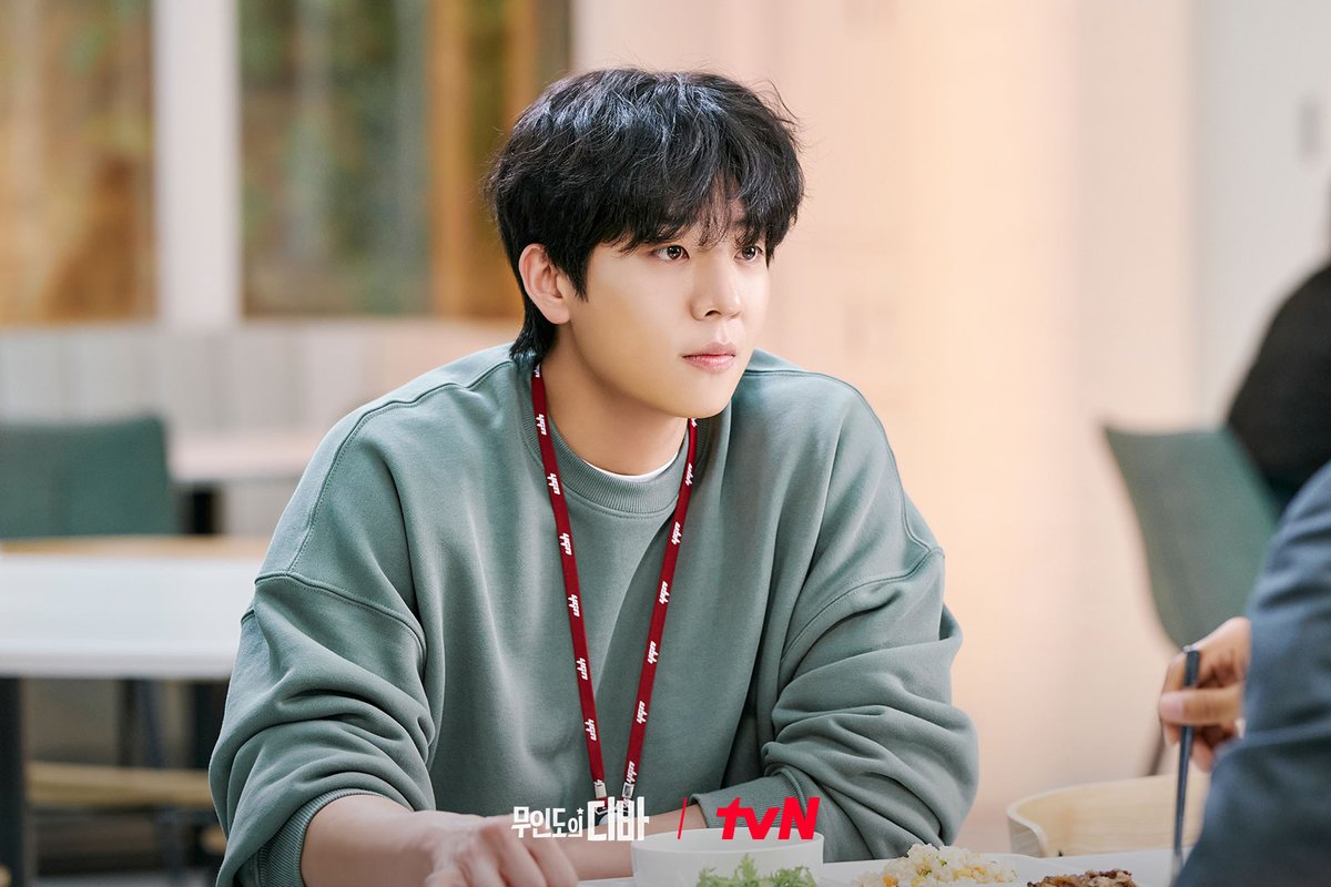 Chae Jong-hyeop como Kang Bo-geol, de moletom verde e crachá pendurado num cordão vermelho, sentado a uma mesa num refeitório