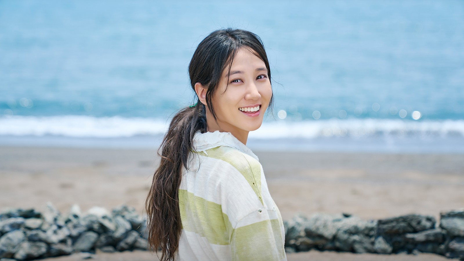 Park Eun-bin como Seo Mok-ha, de rabo de cavalo e blusa listrada em tons de verde, sorri olhando para trás numa praia com mar azul e pedras escuras