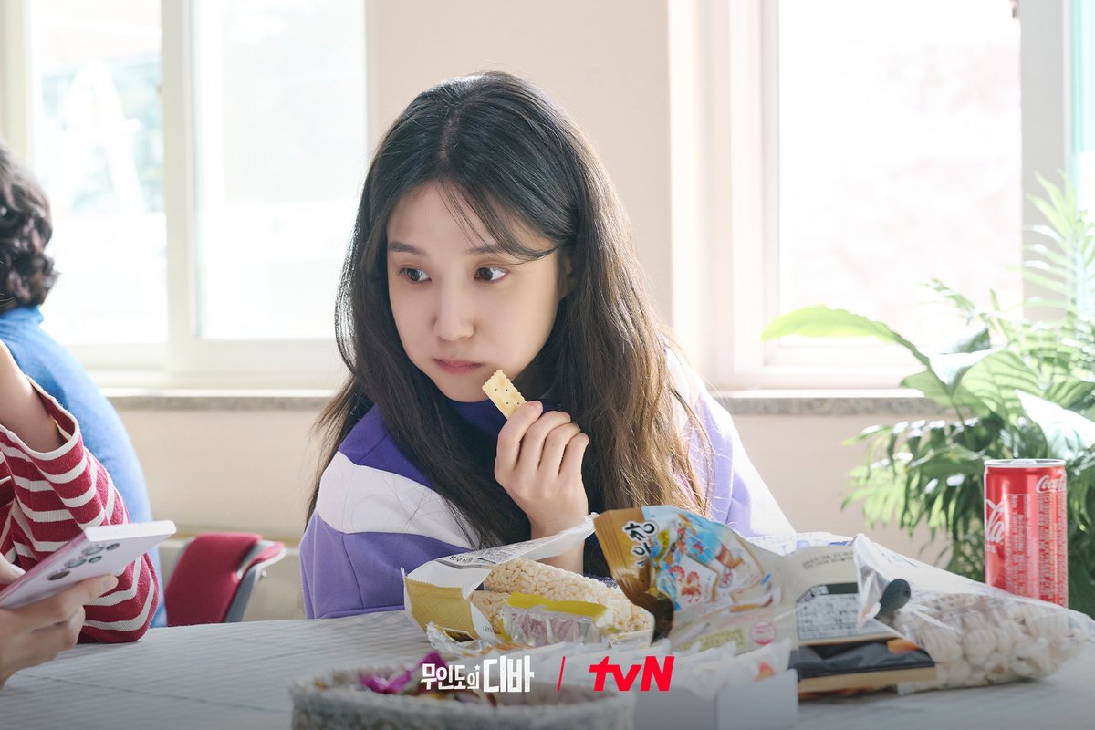 Park Eun-bin como Mok-ha, de blusa listrada roxa e branca, come um salgadinho sentada a uma mesa com pacotes abertos e uma lata vermelha