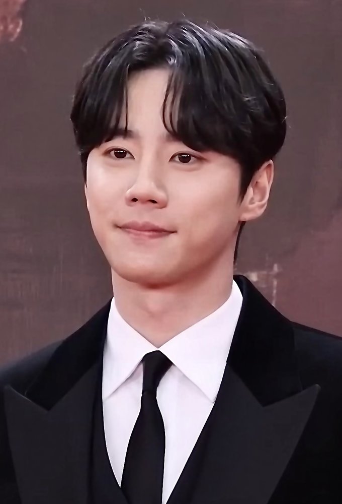 Lee Jun-young, de smoking preto e gravata, sorri de leve diante de fundo escuro em premiação