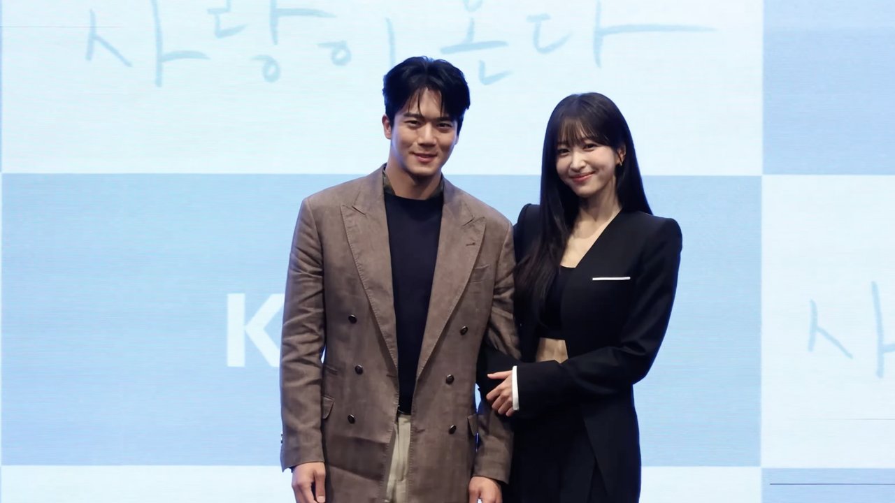 Ha Seok-jin, de paletó marrom, e Hani, de terno preto, posam lado a lado diante do painel azul da coletiva de Tempero do Amor