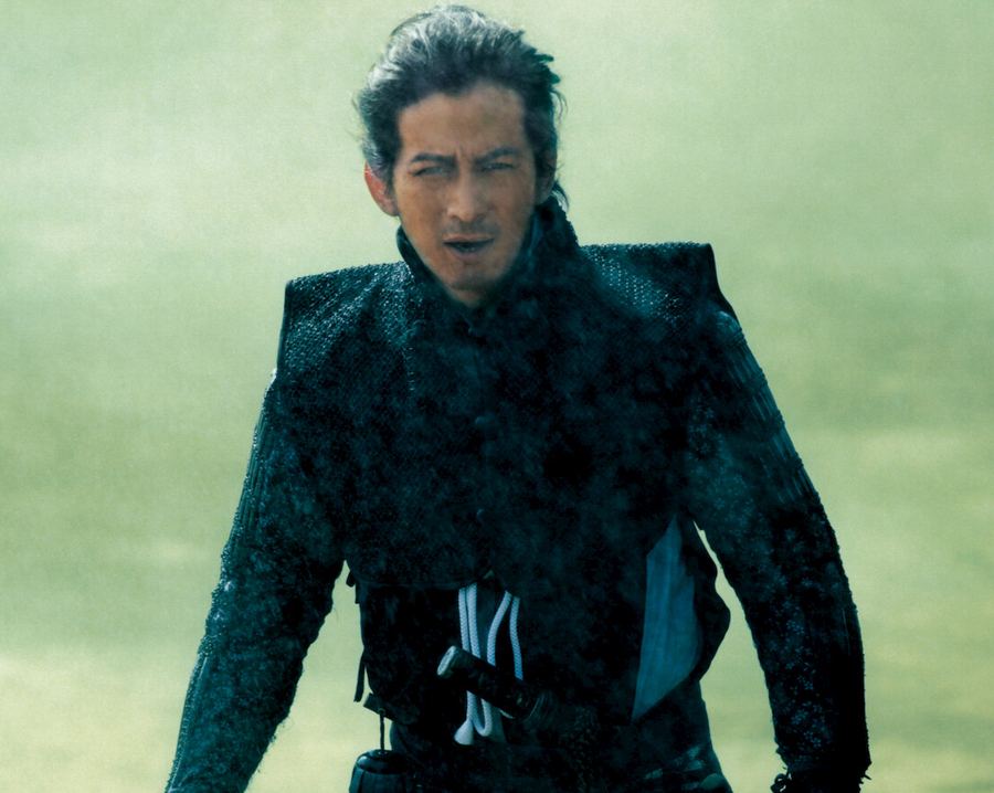 Junichi Okada de armadura escura de samurai, caminhando num campo enevoado