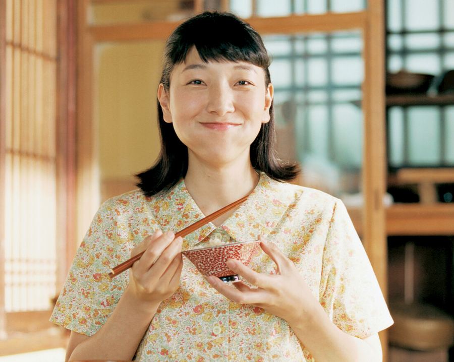 Sakura Ando sorrindo com uma tigela e palitinhos na mão, numa cozinha japonesa de madeira