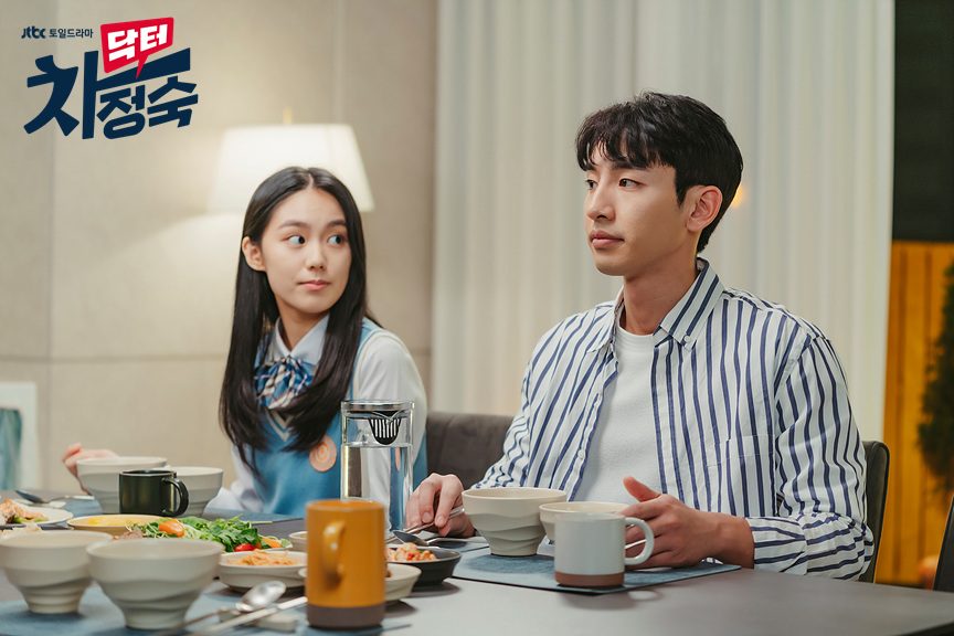 Lee Seo-yeon, de uniforme escolar, e Song Ji-ho, de camisa listrada, à mesa de jantar com tigelas e pratos de acompanhamentos