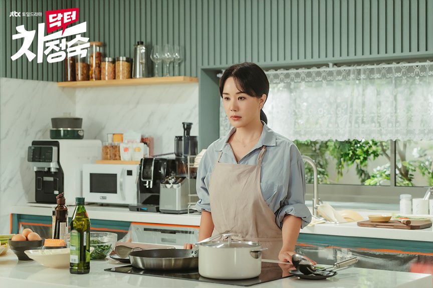 Uhm Jung-hwa como Cha Jeong-suk, de camisa azul-clara e avental bege, séria diante do fogão numa cozinha com panelas, azeite e micro-ondas