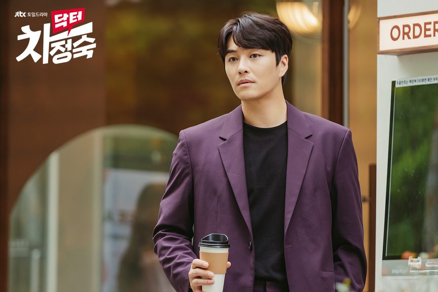 Min Woo-hyuk como Roy Kim, de paletó roxo e camiseta preta, segurando um copo de café para viagem dentro de uma cafeteria