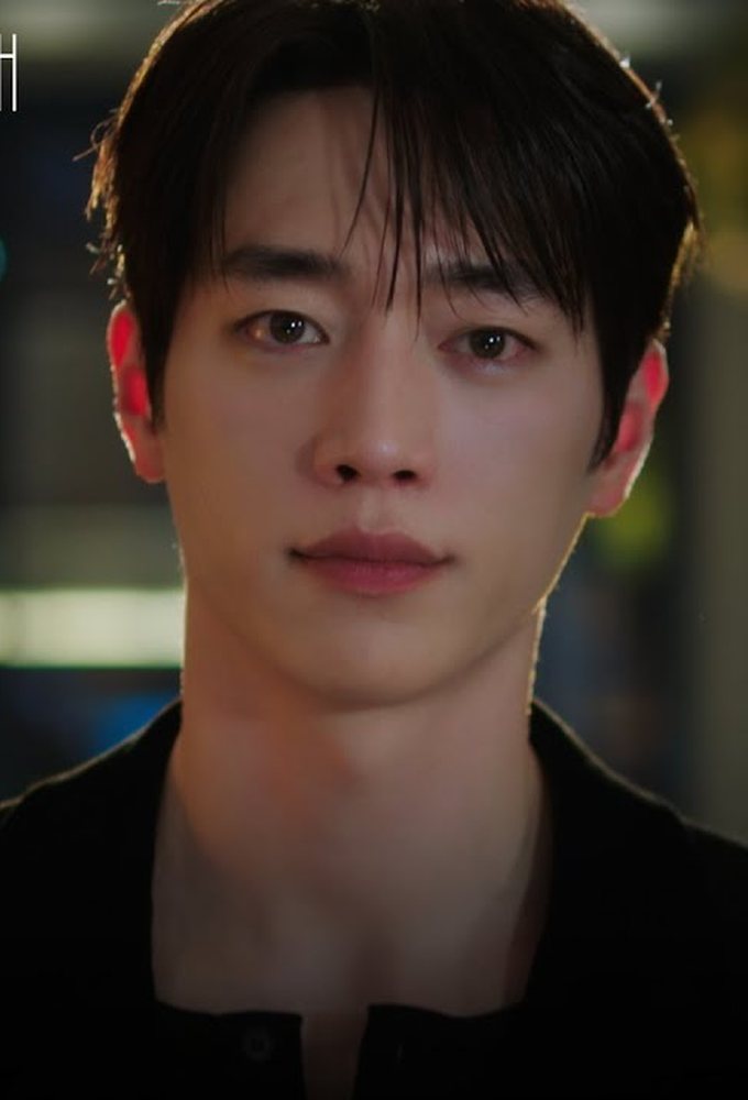 Seo Kang-joon de camisa preta encara a câmera com luzes desfocadas ao fundo, à noite