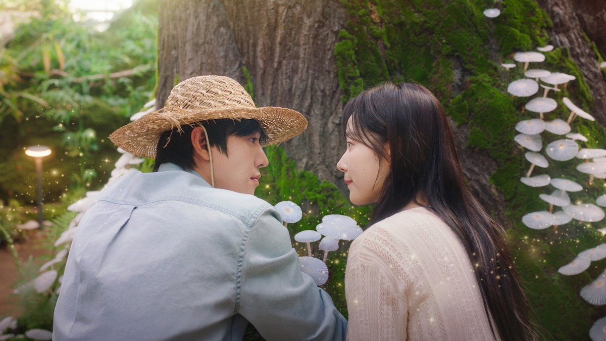 Ahn Hyo-seop de chapéu de palha e camisa azul-clara e Chae Won-been de blusa creme se olham de perfil diante de um tronco com musgo e cogumelos brancos