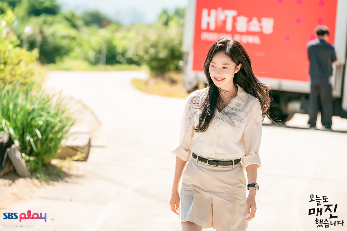 Chae Won-been como Dam Ye-jin, de blusa clara e saia bege, sorri numa estrada do interior com um caminhão vermelho da HIT Homeshopping ao fundo