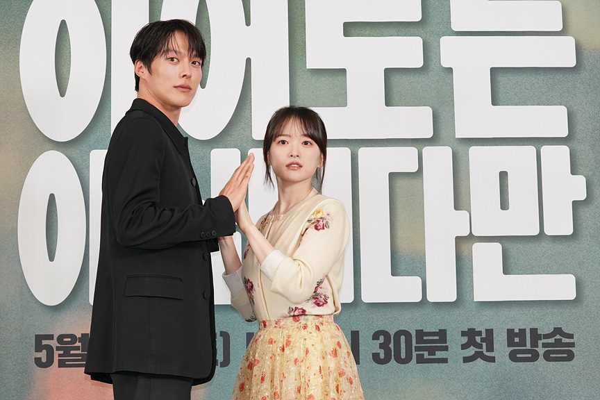 Jang Ki-yong, de terno preto, e Chun Woo-hee, de blusa clara e saia florida, encostam as palmas das mãos diante de um painel com o título coreano da série e o horário de estreia