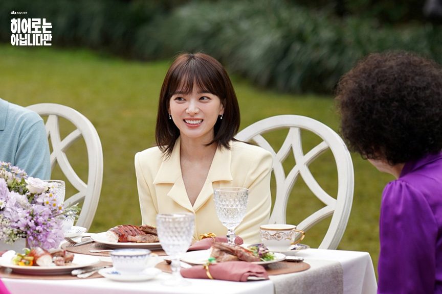 Chun Woo-hee como Do Da-hae, de blazer creme, sorri sentada a uma mesa posta no jardim, diante de uma mulher de cabelo cacheado e blusa roxa vista de costas