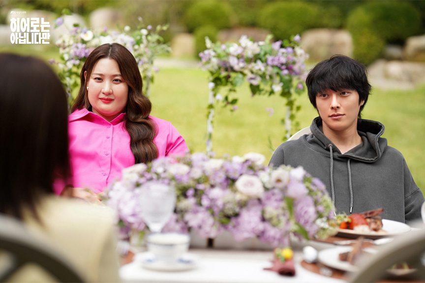Claudia Kim como Bok Dong-hee, de camisa rosa, e Jang Ki-yong como Bok Gwi-ju, de moletom cinza com capuz, sentados a uma mesa com arranjos de flores lilases num jardim