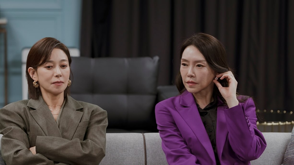 Jang Young Nam e Cha Ji Yeon sentadas num sofá cinza: uma de terno cáqui e braços cruzados, a outra de terno roxo tocando a orelha