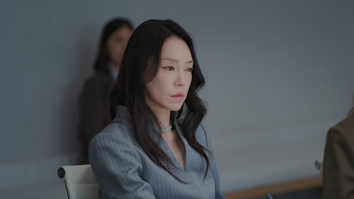 Cha Ji Yeon de blusa cinza, séria, sentada à mesa de reunião com um lápis na mão