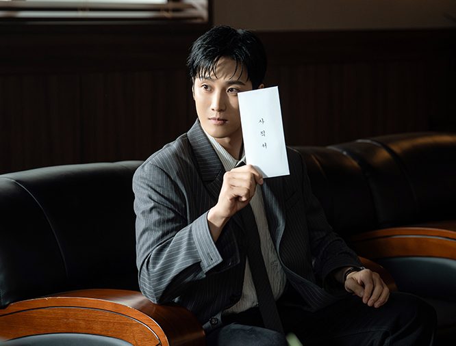 Ahn Bo-hyun de terno risca de giz sentado num sofá de couro segura um envelope branco escrito em coreano