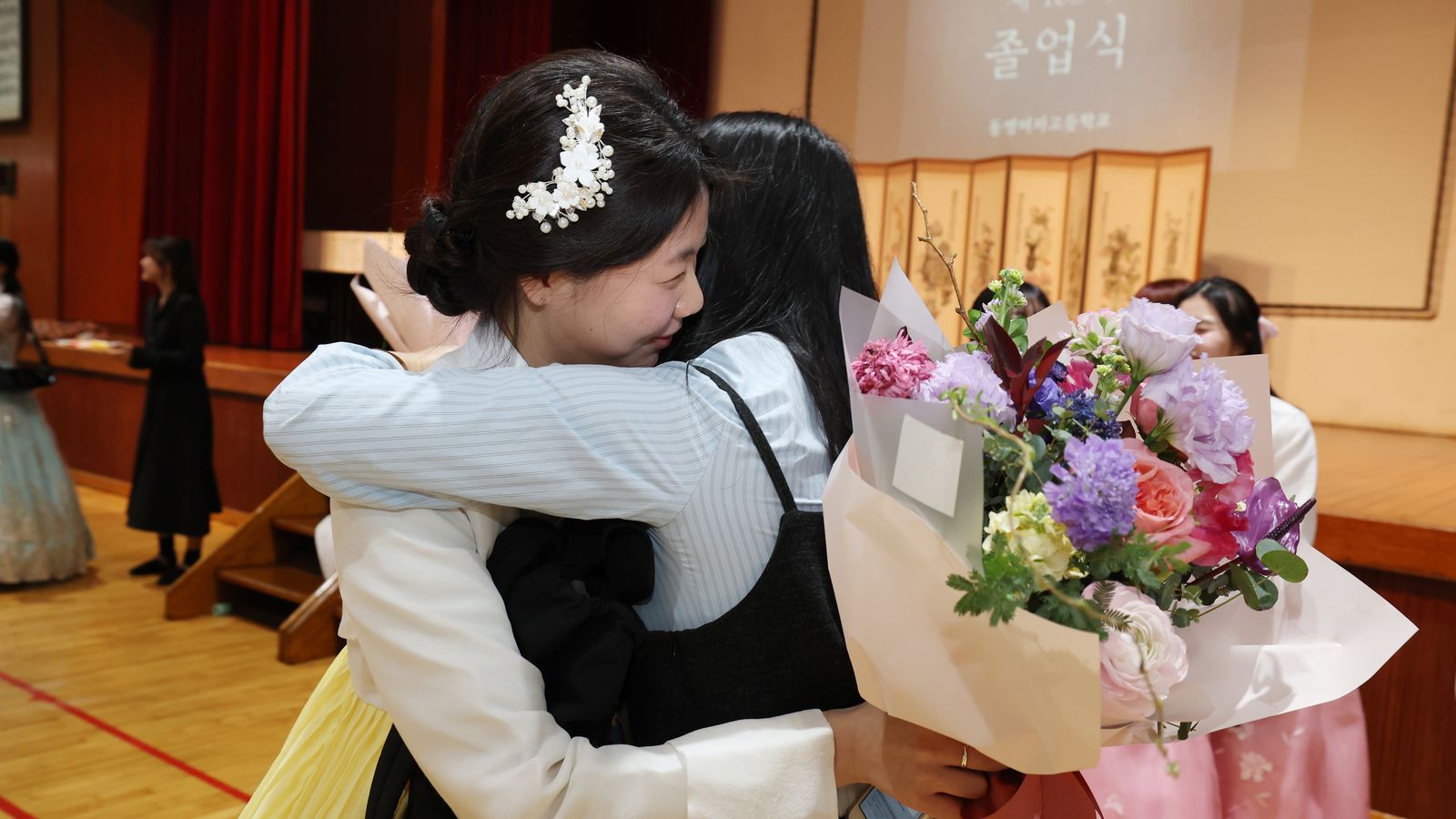 Formanda de hanbok com enfeite branco no cabelo sorri de olhos fechados enquanto é abraçada por uma colega que segura um buquê de flores, diante de um palco