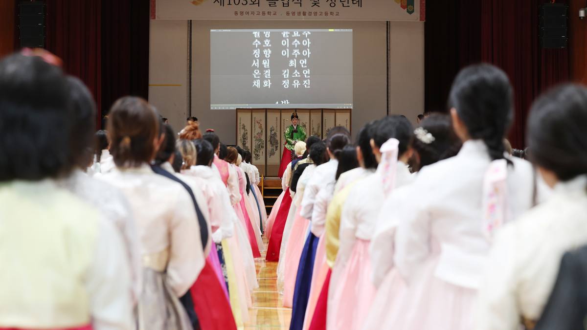 Fila de formandas de hanbok vistas de costas caminhando em direção ao palco, onde uma tela mostra nomes em coreano