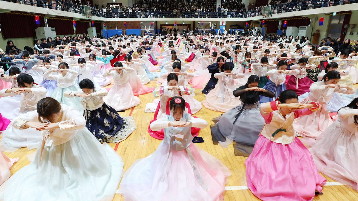 Dezenas de formandas de hanbok colorido ajoelhadas no piso de um ginásio, com as mãos unidas na testa, fazendo reverência