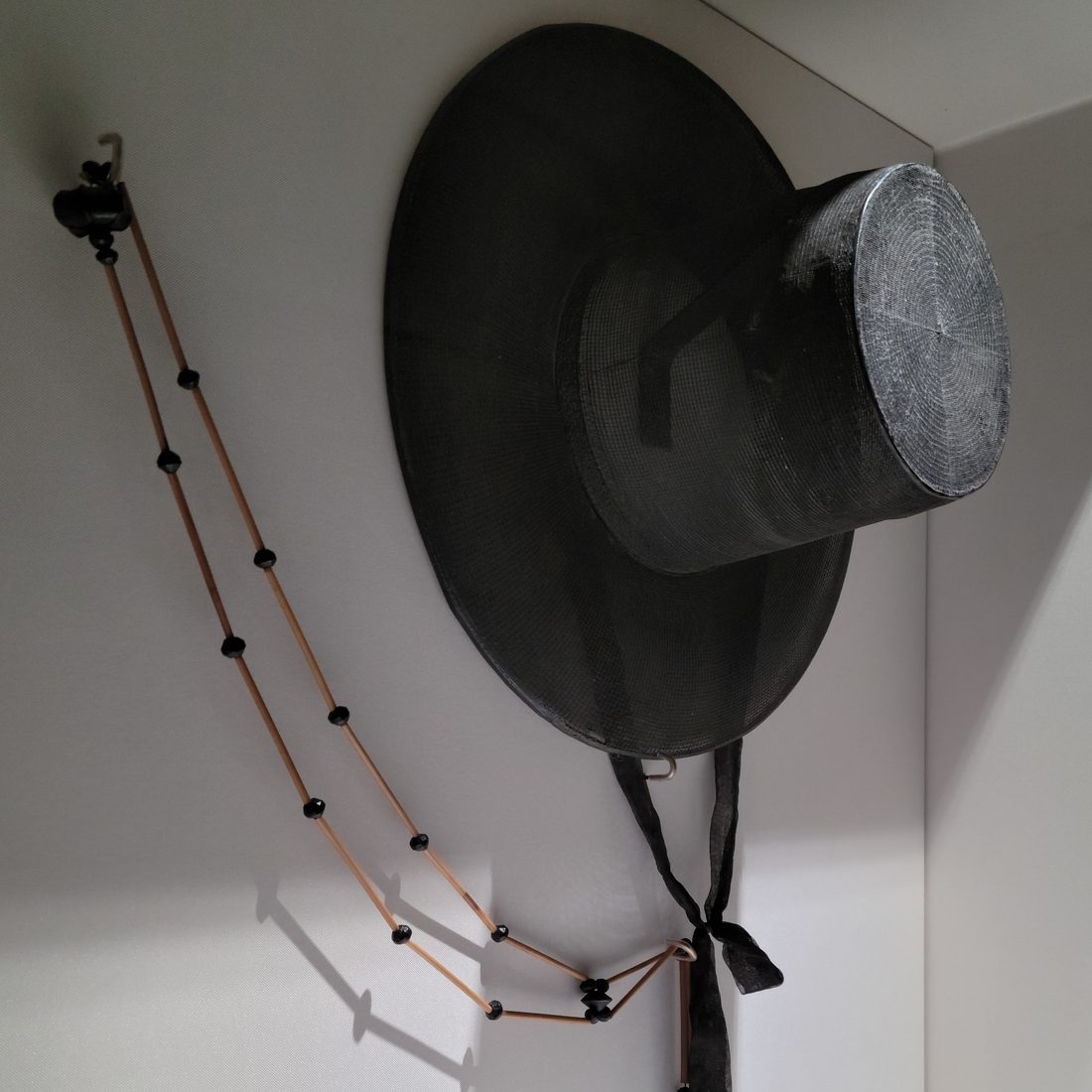Gat preto de aba larga e copa cilíndrica, com um cordão de segmentos bege e contas pretas pendurado ao lado e fita preta