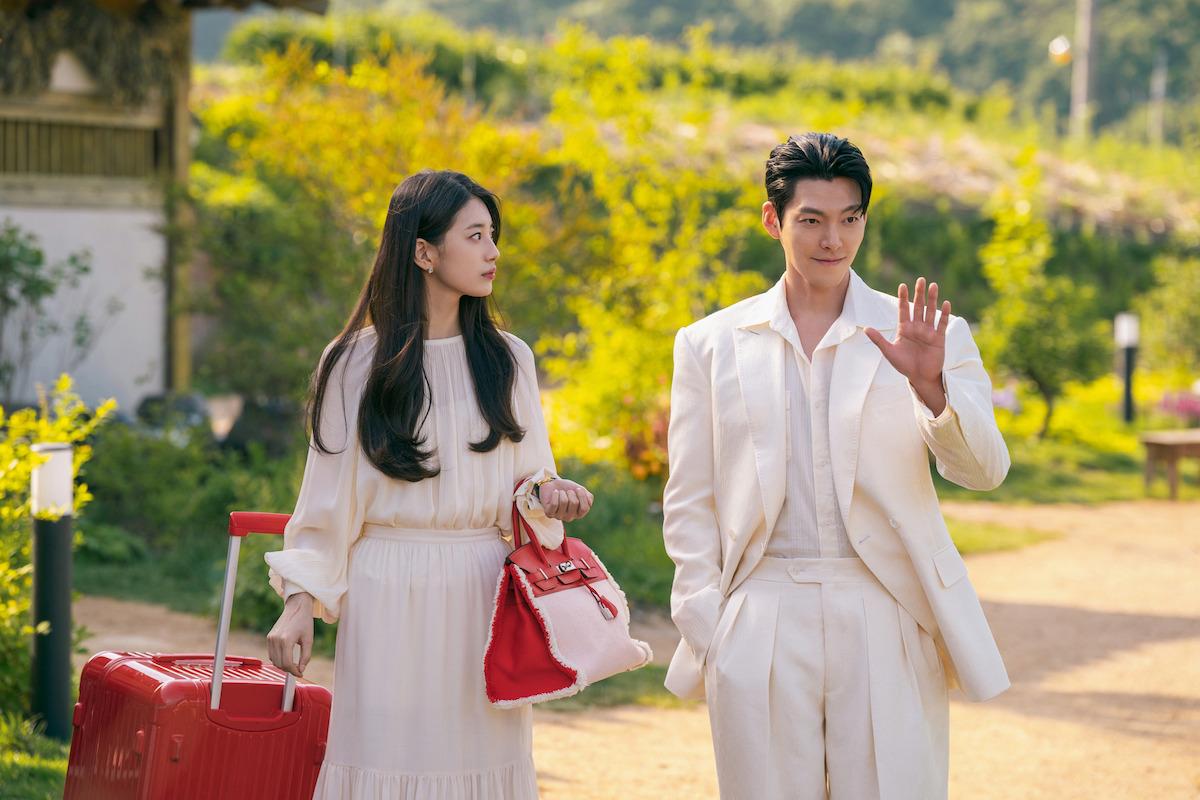 Suzy como Ka-young, de vestido branco com bolsa e mala vermelhas, olha para Kim Woo-bin como Iblis, de terno branco, que acena numa rua arborizada