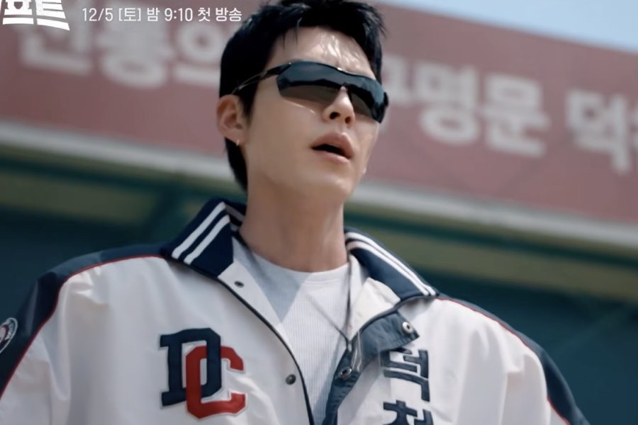 Kim Woo-bin de óculos escuros e agasalho branco e azul com as letras DC, diante de um muro vermelho com letras coreanas, e a data 12/5 no alto