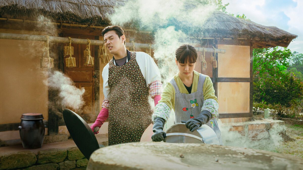 Kang Ha-neul de avental florido e luvas cor-de-rosa e Ko Min-si de avental xadrez e luvas pretas destampando caldeirões fumegantes diante de uma casa de teto de palha