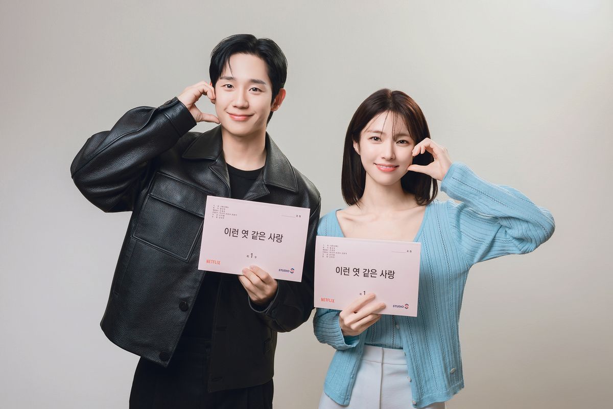 Jung Hae-in, de jaqueta de couro preta, e Ha Young, de cardigã azul-claro, fazem corações com as mãos segurando roteiros com o título coreano 이런 엿 같은 사랑