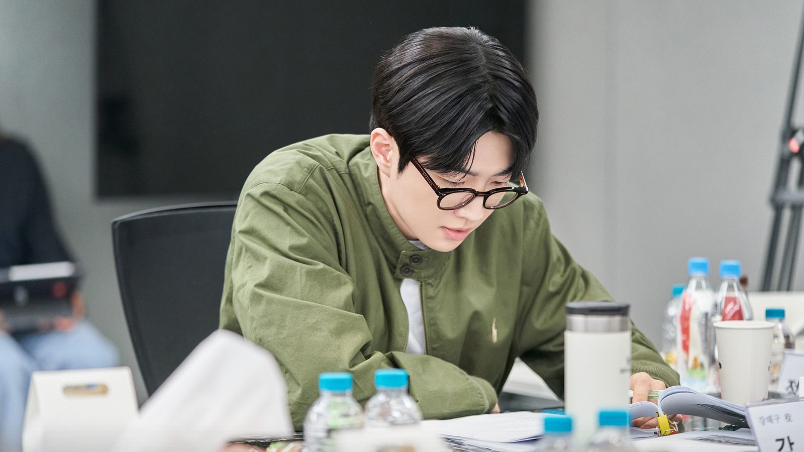 Kang Hoon de óculos e jaqueta verde, debruçado sobre o roteiro durante a leitura de Happy Anding