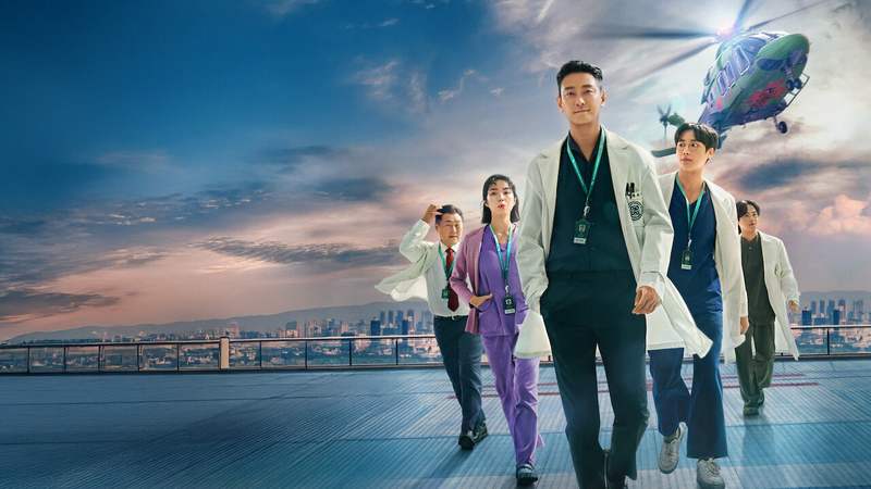 Ju Ji-hoon de jaleco branco à frente de quatro colegas num heliponto, com um helicóptero ao fundo