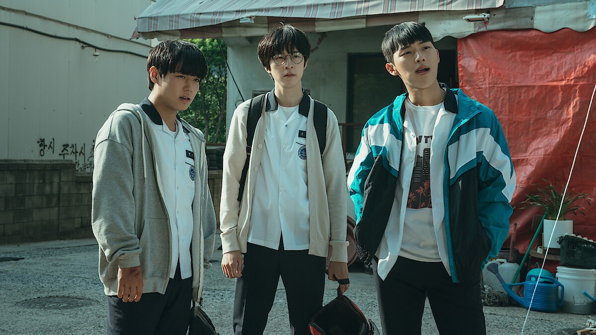 Os três protagonistas da Classe 1, Park Ji-hoon, Choi Hyun-wook e Hong Kyung, de uniforme escolar numa rua, um de moletom cinza, outro de óculos e mochila e o terceiro de jaqueta esportiva