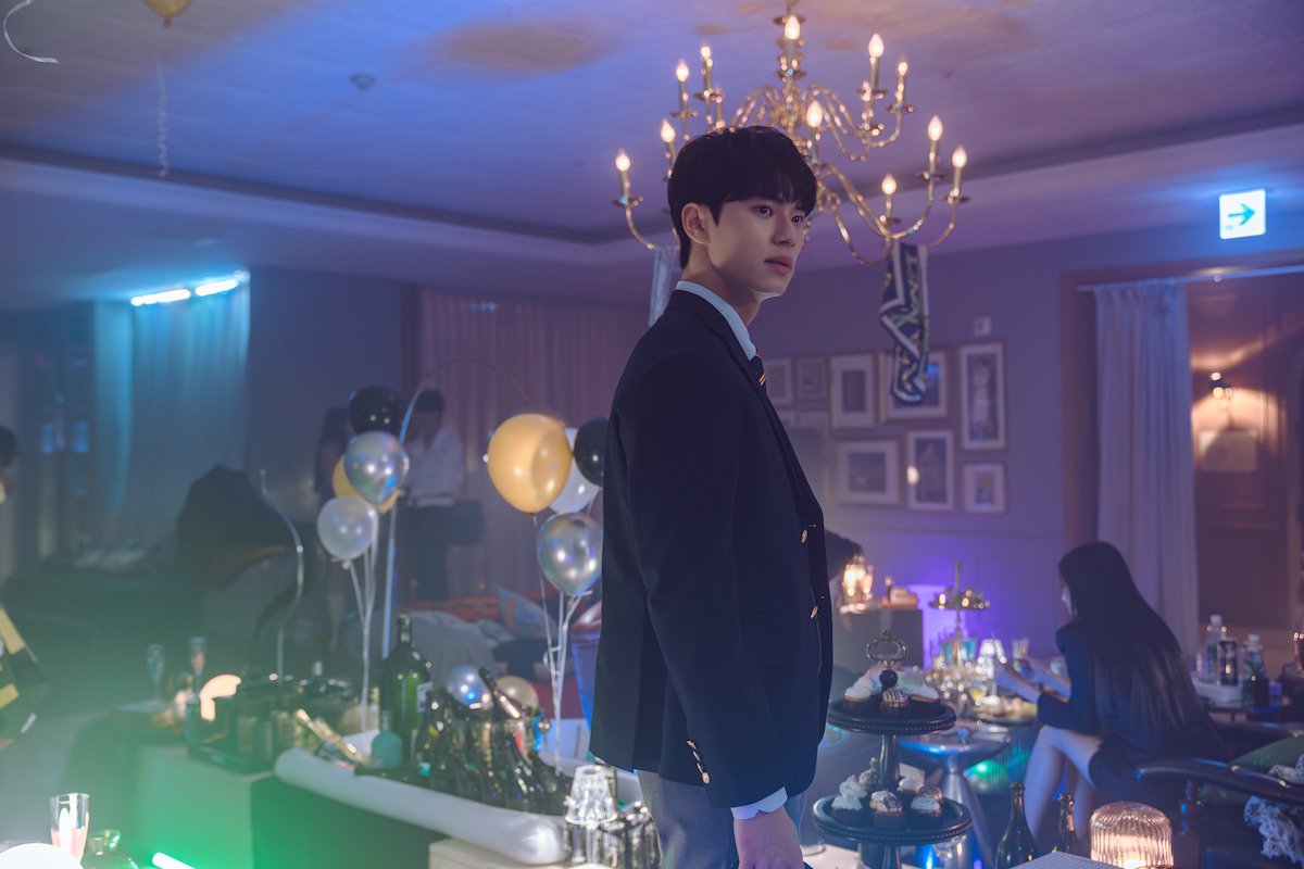 Lee Chae-min como Kang Ha, de uniforme escolar, de pé no meio de uma festa em salão com lustre, balões e luzes coloridas