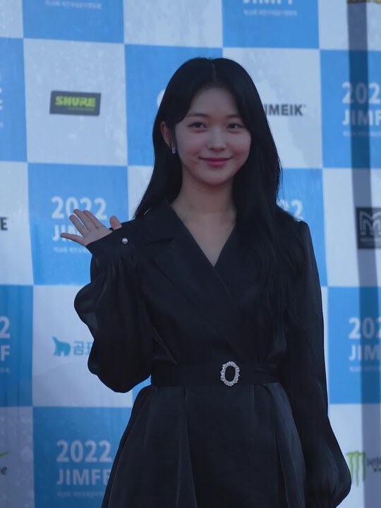 Moça de cabelo preto longo e liso, sorrindo e acenando com a mão direita, de vestido preto com cinto de fivela brilhante, diante de um painel azul e branco de patrocinadores