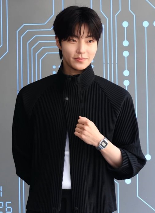 Hwang In-youp de jaqueta preta plissada e relógio no pulso, posando diante de um painel com desenho de circuito