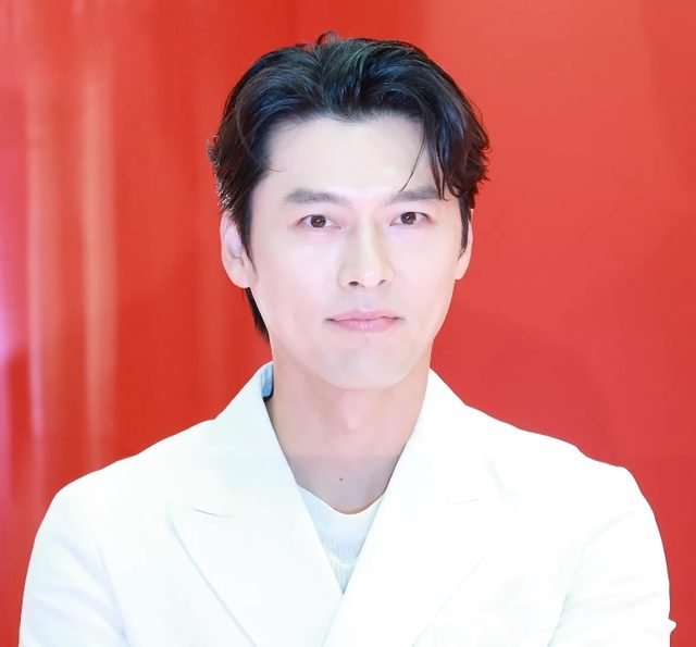 Hyun Bin de blazer branco diante de fundo vermelho