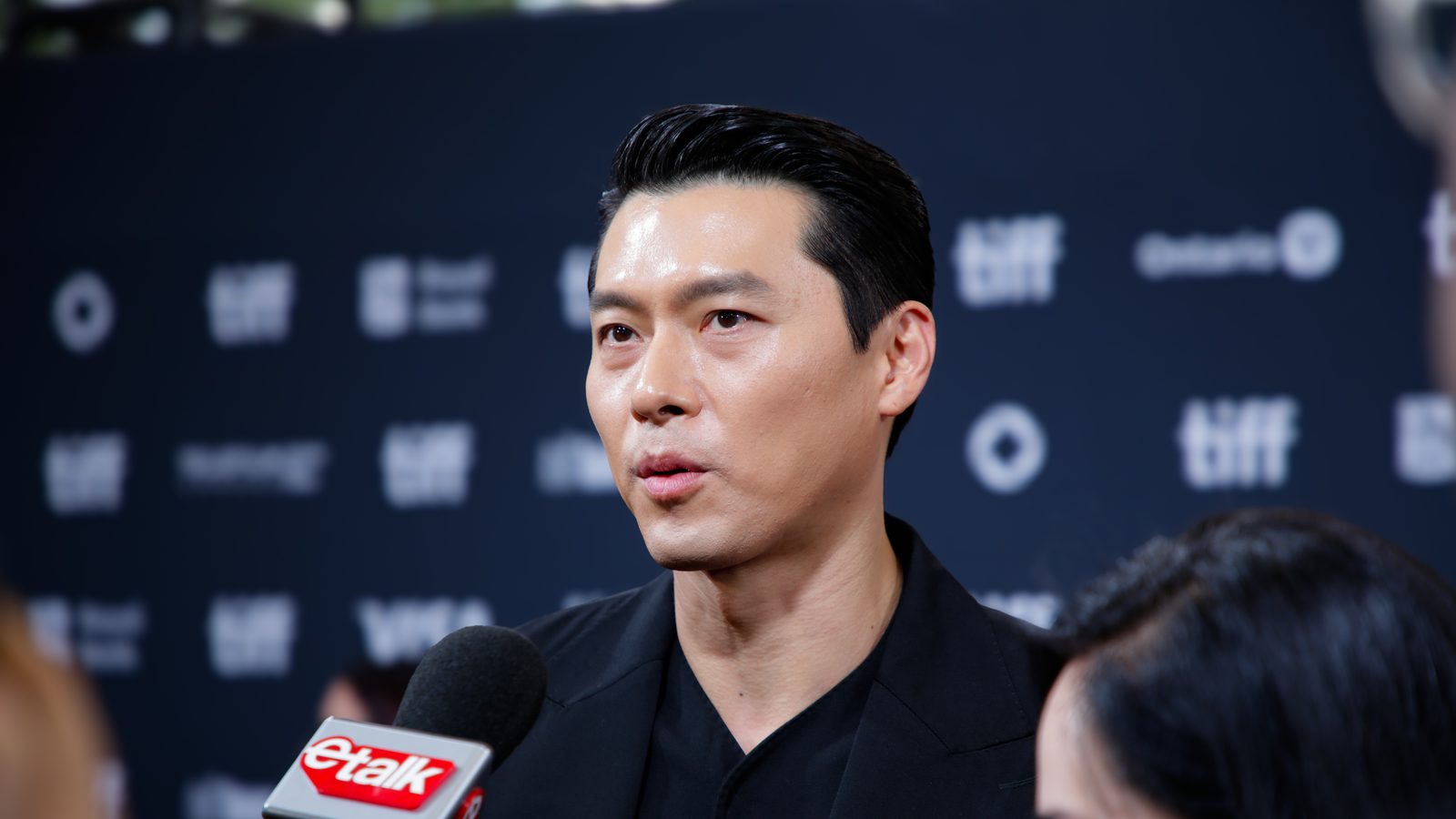 Hyun Bin de terno preto dá entrevista a um microfone diante de painel do Festival de Toronto
