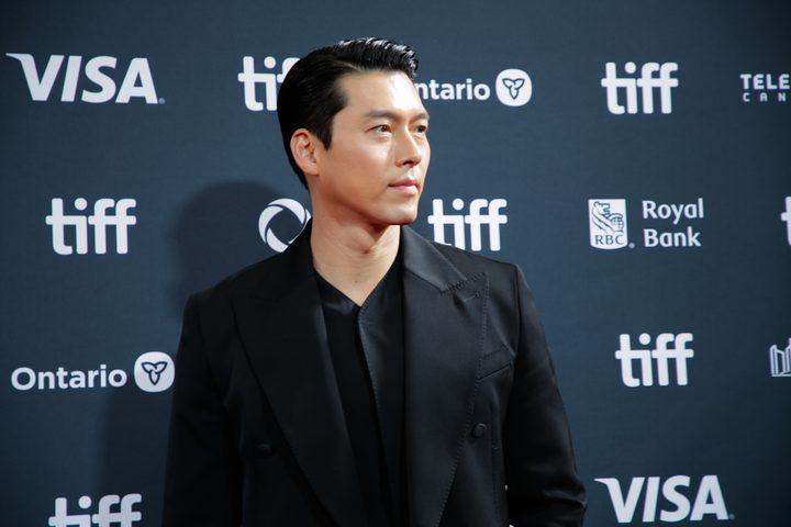 Hyun Bin de terno preto olhando para o lado diante de painel com logotipos do festival