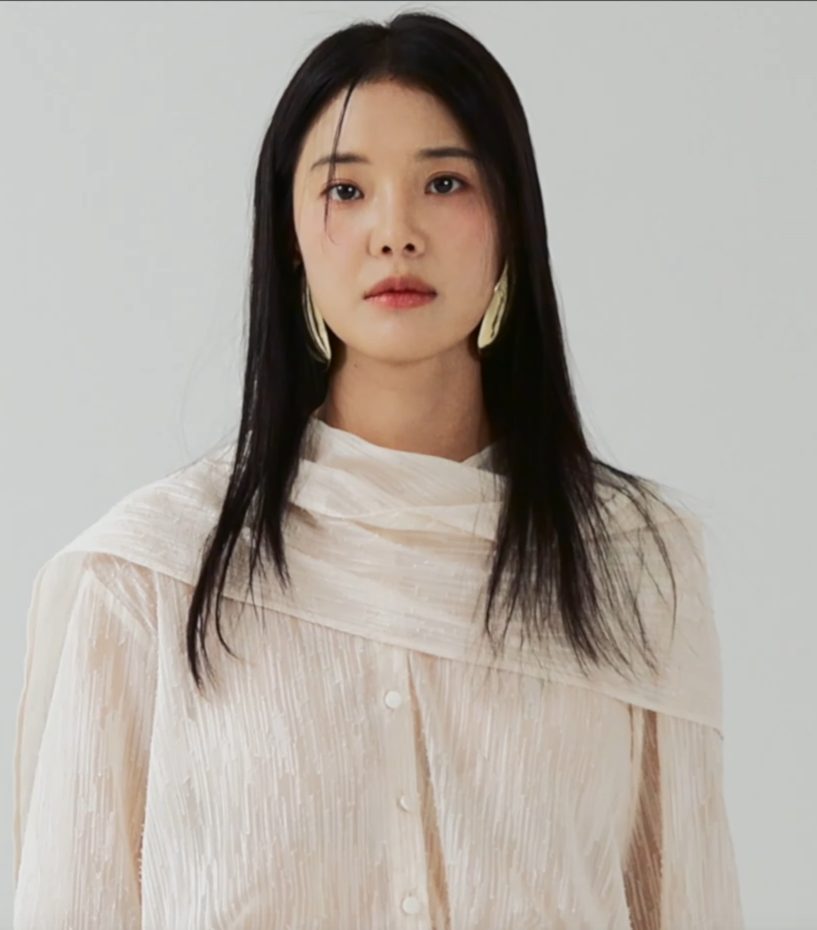 Mulher de cabelo preto liso e comprido, brincos dourados grandes e blusa creme com lenço no pescoço, diante de um fundo cinza claro