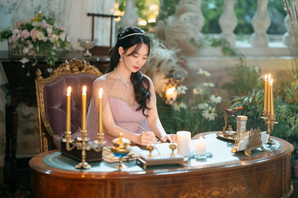 Mulher de cabelo preto longo, com tiara fina e vestido rosado bordado, escrevendo sentada numa escrivaninha antiga cercada de velas acesas e flores brancas