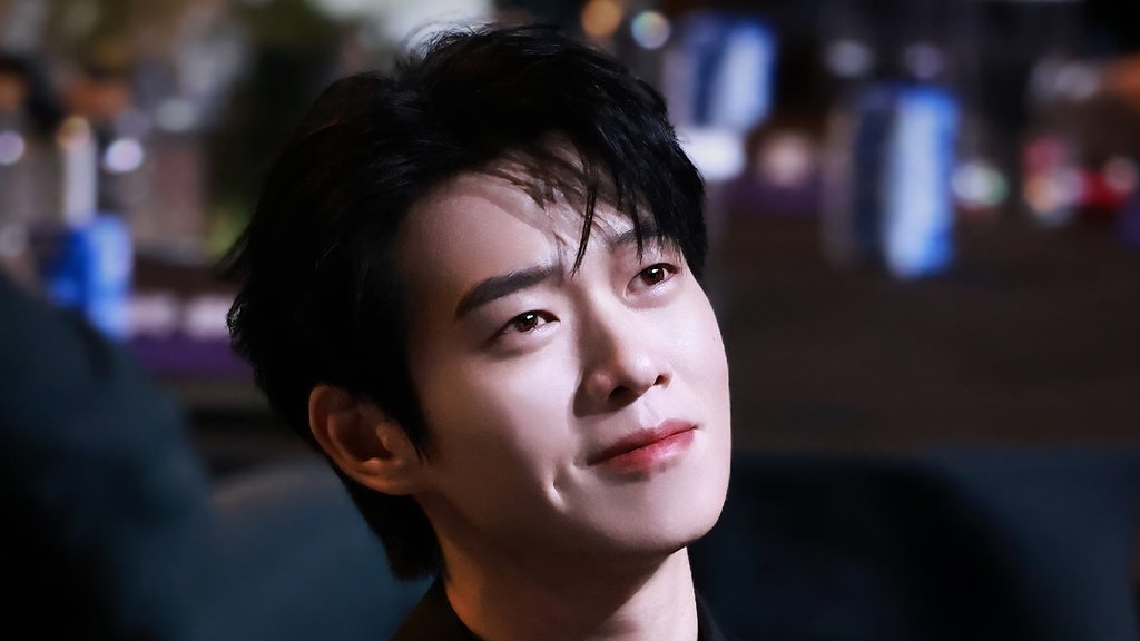 Wei Daxun de terno e camisa pretos, sentado, sorrindo e olhando para cima, com luzes desfocadas ao fundo