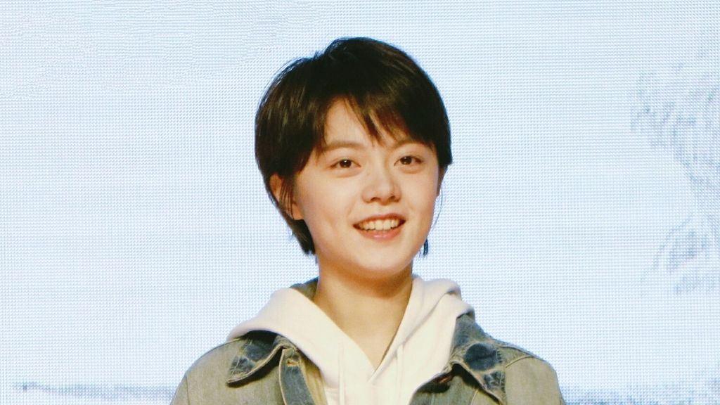 Zhao Jinmai de cabelo curto, moletom claro e jaqueta jeans, sorrindo diante de um painel azul claro