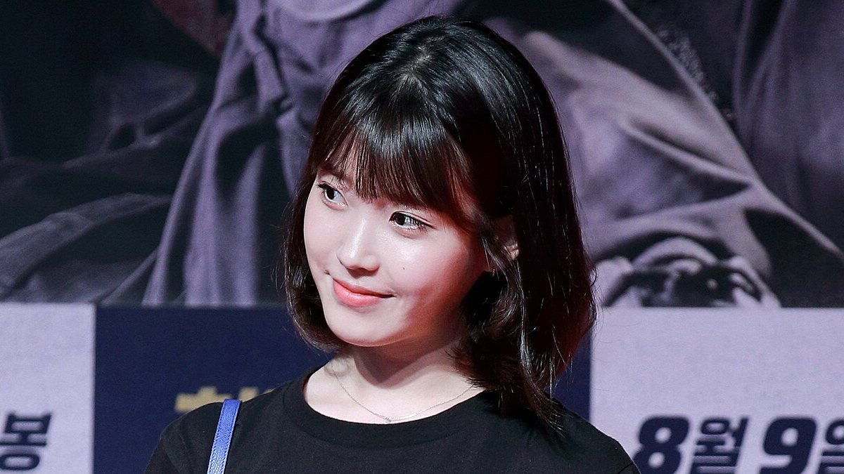 IU de cabelo curto com franja e camiseta preta sorri olhando para o lado diante de um painel de divulgação