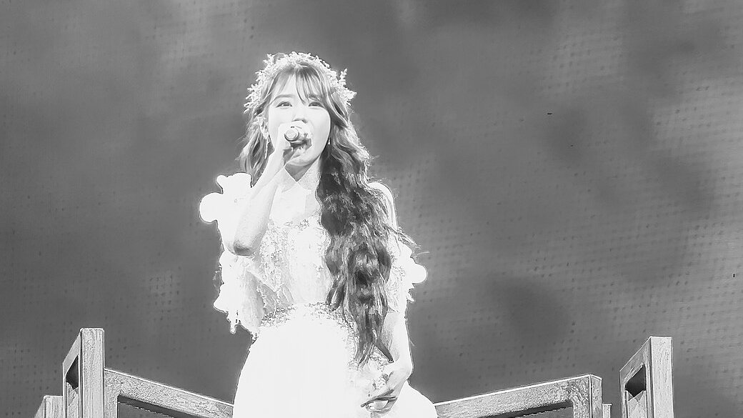 Foto em preto e branco de IU de vestido branco e coroa de flores cantando ao microfone no alto de um palco