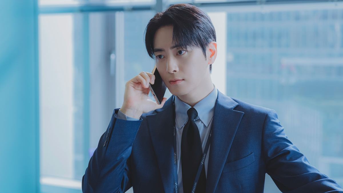 Homem de cabelo preto com franja, terno azul-marinho, camisa azul-clara e gravata escura, falando ao celular diante de uma janela de escritório
