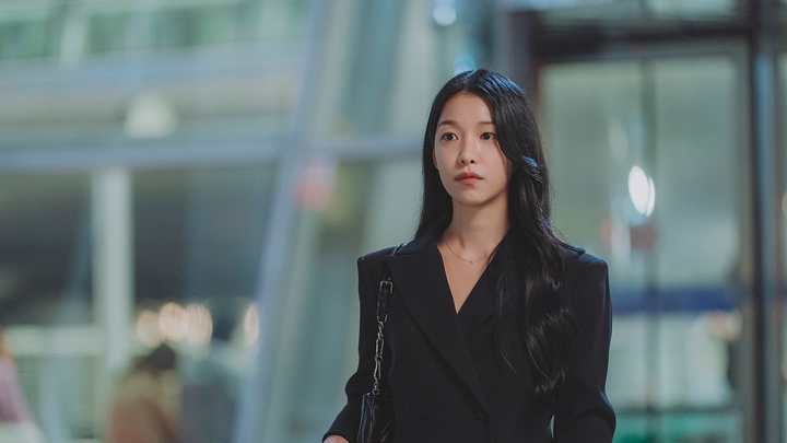Mulher de cabelo preto longo e blazer preto, com bolsa de corrente no ombro, caminhando num saguão envidraçado