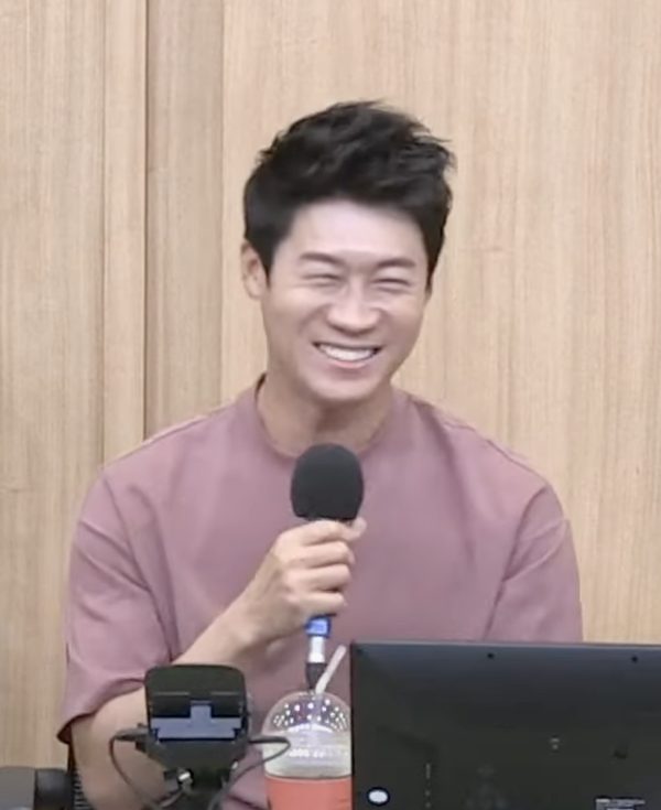 Homem de cabelo preto curto, sorrindo de olhos fechados, de camiseta rosa, segurando um microfone preto num estúdio de rádio com parede de madeira clara